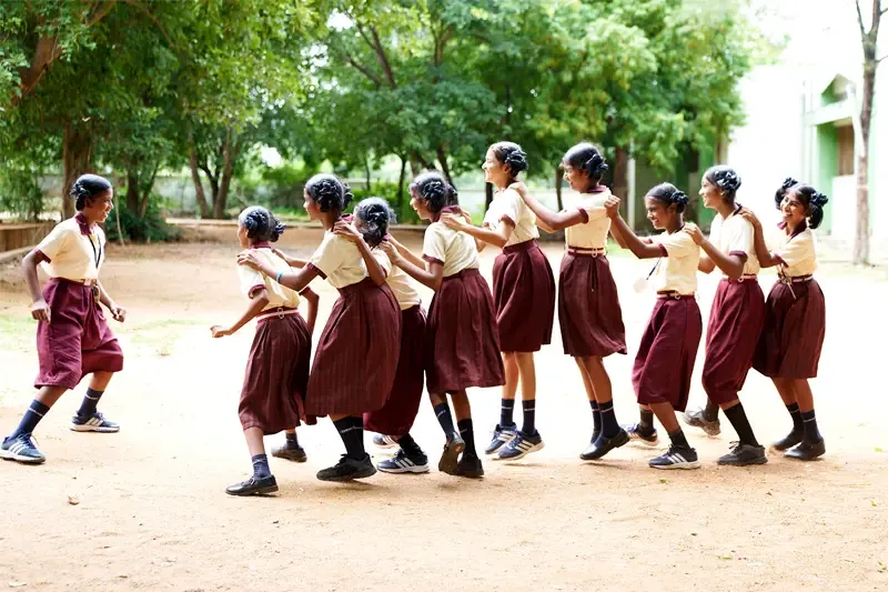 A line of Indiana girls in matching tan and maroon school uniforms hold each other's shoulders and playfully skip across a dirt schoolyard. Trees and a school building can be seen in the background.